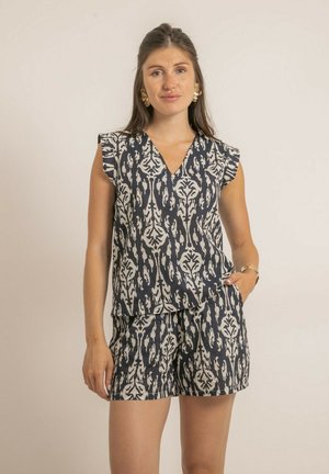 Femme portant un ensemble assorti de haut sans manches et short à motifs bleu marine et beige, debout avec une main dans la poche devant un fond uni.
