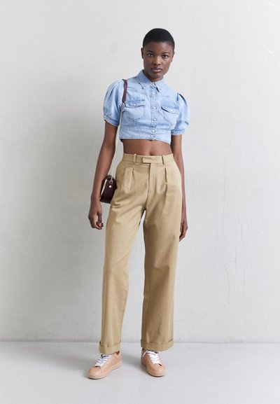 Mujer vistiendo una camisa de mezclilla corta azul clara, pantalones de talle alto beige, zapatillas melocotón y llevando un pequeño bolso de hombro oscuro.