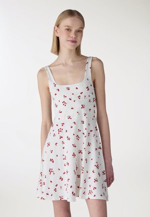 Giovane donna che indossa un vestito bianco senza maniche con motivo di ciliegie rosse, in piedi e leggermente sorridente su uno sfondo semplice.