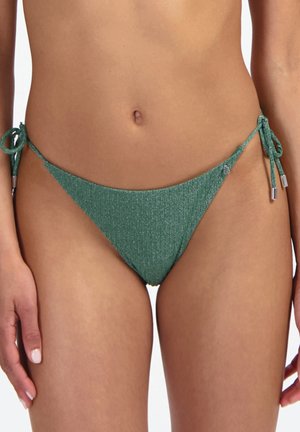 Slip bikini verde a coste con lacci laterali indossato da una persona in piedi, che mostra l'addome e la parte superiore delle cosce.