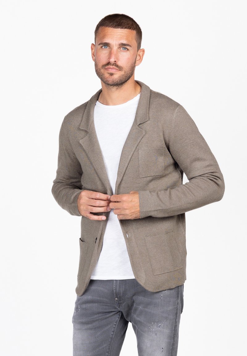 Beige cardigan met een reverskraag, sluiting met één knoop en twee voorzakken, gedragen over een wit shirt, gecombineerd met grijze denim.