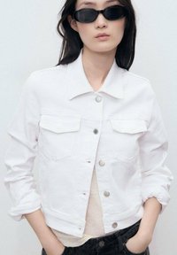 White denim jacket with two chest pockets, silver buttons, and a collared neckline. Textured fabric, cropped design, and rolled sleeves.