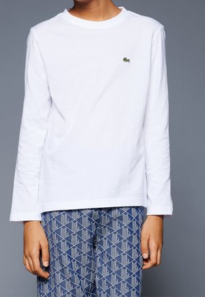 Osoba ubrana w białą koszulę z długim rękawem z małym zielonym logo krokodyla oraz niebieskie spodnie we wzory na jednolitym szarym tle.