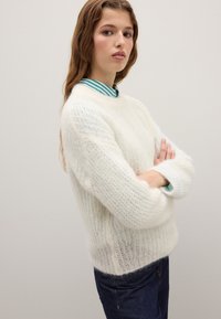 Cream-colored knitted sweater with a soft, textured finish. Features long sleeves with ribbed cuffs. Worn over a striped shirt.