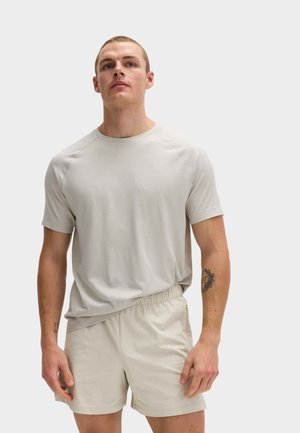 Ung mand iført lysebeige kortærmet skjorte og matchende shorts med elastisk talje, stående mod en ensfarvet hvid baggrund.