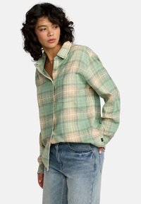 Camicia a quadri verde e beige con colletto, maniche lunghe, vestibilità rilassata e texture morbida, abbinata a jeans in denim blu.
