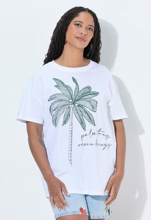 Femme portant un t-shirt blanc avec un dessin de palmier et du texte, associé à un short en denim bleu clair avec de petites patches fruits.