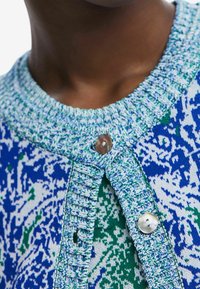 Neulettu neuletakki, jossa on sinivihreä kuvio, pyöreä pääntie teksturoidulla reunuksella ja helminappisuljenta.