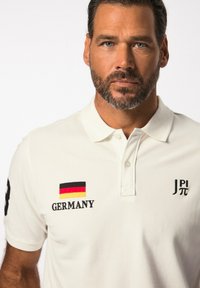 Bílá polo košile vyrobená z bavlny, s černými a červenými detaily, výšivkou "NĚMECKO", nášivkou vlajky a logem na hrudi. Klasický střih.