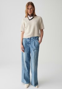 Vrouw in een kort mouw cremekleurige trui, met een patroon op de halsdoek, wijde lichtblauwe jeans en witte puntige schoenen, staande met één hand in de zak.