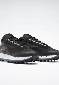 Paire de baskets noires de sport avec mesh texturé, lacets noirs et semelle blanche rembourrée avec des crampons pour l’adhérence.
