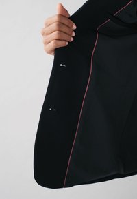 Sort blazer med en glat tekstur, kontrastfarvet pink piping langs kanterne og to synlige metallukninger indeni foret.