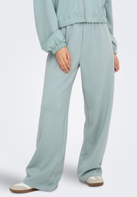 Pantalon large taille haute vert clair en tissu doux avec poches latérales et taille élastique, assorti de chaussures beiges à enfiler.