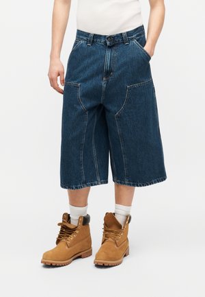 Personne portant un short en denim sombre ample arrivant aux genoux, des chaussettes blanches et des bottes à lacets couleur beige, debout devant un fond uni.