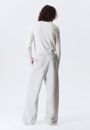 Personne aux cheveux bouclés portant une chemise blanche à manches longues et un pantalon large gris clair, debout avec les mains dans les poches arrière.