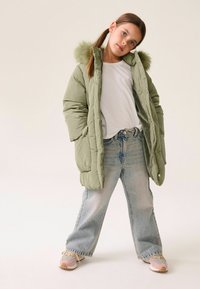 Olivegrüner Pufferjacke mit einer Kapuze aus Kunstfell, kombiniert mit hellblauen, weiten Jeans und mehrfarbigen Turnschuhen. Darunter ein schlichtes weißes T-Shirt.