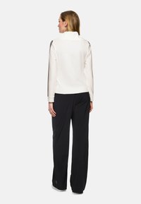 Weißer Reißverschluss-Pullover mit gerippter Textur und Funnel-Neck, kombiniert mit lockeren schwarzen Hosen mit seitlichen Streifen.