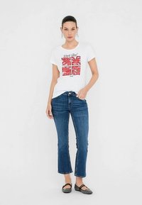 Balti marškinėliai su spalvotu grafiniu atspaudu, vaizduojančiu raudonas lūpas, derinami su mėlynomis tiesiomis džinsų kelnėmis ir juodais butais. Paprastas dizainas.