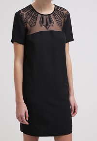 Černé šaty s krátkým rukávem, které mají průhledný vyšívaný výstřih. Tkanina je hladká, s přiléhavým střihem a přímým střihem.