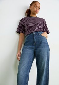 Camiseta de gran tamaño a rayas en púrpura y negro combinada con vaqueros de tiro alto y pierna ancha en denim azul. Textura suave, ajuste casual y con un mínimo de elementos metálicos.