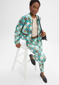 Zweiteiliges Outfit mit einer jacke im Argyle-Muster in Türkis, Braun und Beige, dazu passende Hose. Schwarze Slipper und eine kleine Tasche.