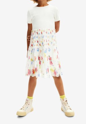 Robe blanche à manches courtes avec une jupe en couches transparente ornée d'imprimés floraux colorés ; associée à des baskets montantes de couleur claire.