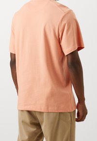 Camiseta de algodón color melocotón claro con mangas cortas, cuello redondo y un detalle de costura sutil, combinada con pantalones color beige, vista desde la parte de atrás.