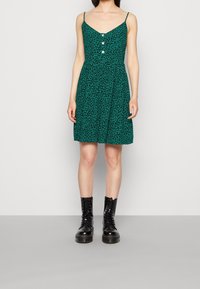 Vestido verde con un estampado de leopardo, escote en V, botones en la parte delantera y tirantes finos, combinado con botas negras con cordones.