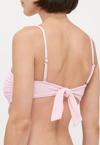 Ružový textúrovaný bikiny top s nastaviteľnými ramienkami a uzlíkovým uzáverom na chrbte; má jemný vzor a hladký povrch látky.