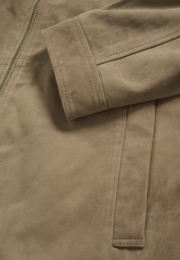 Jachetă din piele întoarsă bej, cu închidere cu fermoar, textură netedă și mânecă bine croită, cu detalii de cusături.