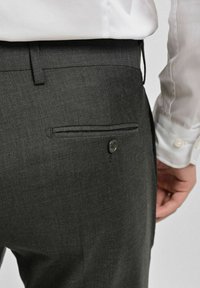 Mörkgrå byxor i texturerat tyg med en bakficka som har knäppning. En vit skjorta är delvis synlig.