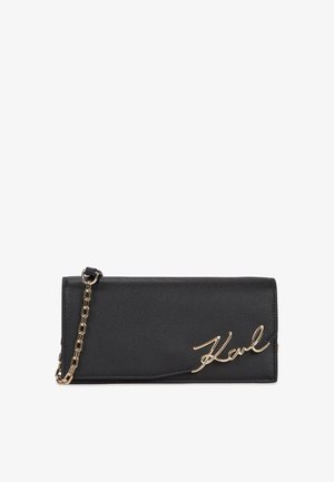 Carteira de couro preto com uma alça de corrente dourada, apresentando o logótipo "Karl" em estilo cursivo na frente, textura suave e design elegante.