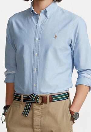 Mand iført lyseblå skjorte med knapper og broderet logo, stripet stofbælte med læderdetalje og beige bukser, hænderne i lommerne.