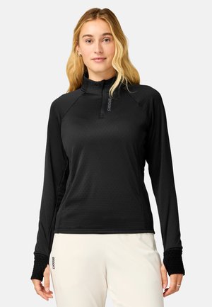 Mujer con cabello rubio largo que lleva un jersey negro con cremallera hasta el cuello y pantalones blancos sobre un fondo liso.