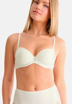 Jonge vrouw met steil blond haar, gekleed in een witte beugel-bh en bijpassend hooguitgesneden ondergoed, tegen een effen achtergrond.