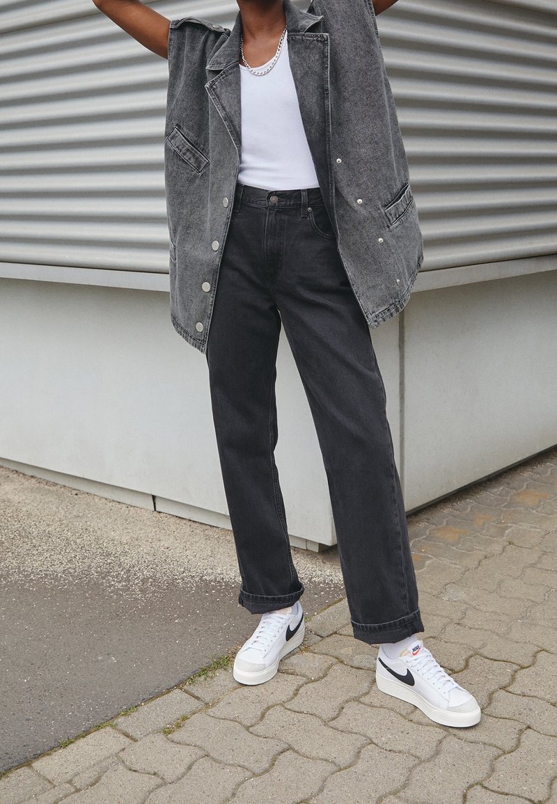 Personne portant une veste en jean grise sans manches, un débardeur blanc, un jean noir et des baskets blanches Nike, debout sur un trottoir pavé.