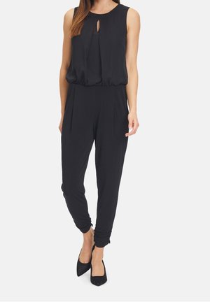 Kvinde iført en ærmeløs sort jumpsuit med nøglehul ved halsen og tilspidsede bukser, kombineret med sorte spidse stiletter.