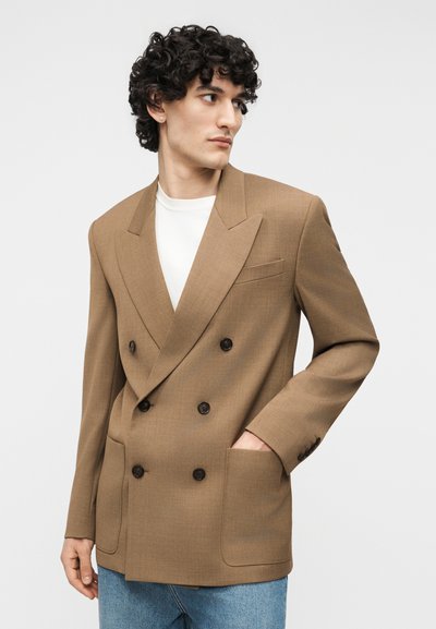 Νέος άνδρας με κατσαρά μαλλιά που φοράει καφέ διπλόσφιγκο σακάκι πάνω από λευκό πουκάμισο και μπλε τζιν, κοιτάζοντας προς τα πλάγια, με το ένα χέρι στην τσέπη.