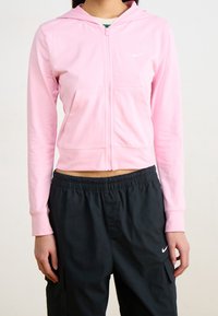 Kvinna som bär en rosa zip-up hoodie och svarta Nike sweatpants, stående med armarna avslappnat längs sidorna mot en enkel bakgrund.