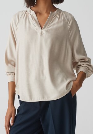 Woman wearing a loose beige long-sleeve V-neck blouse with rolled cuffs and dark navy pants, one hand in pocket.