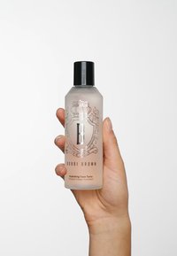 Transparent plastflaska med svart kork, prydd med en silverlogotyp och text för Bobbi Brown Hydrating Face Tonic, hålls i en hand.