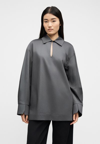 Γυναίκα με μακριά μαύρα μαλλιά, φορώντας υπερμεγέθη γκρι μπλούζα με λαιμόκοψη σε σχήμα κλειδαρότρυπας και φαρδιά μανίκια, σε συνδυασμό με μαύρο παντελόνι, στέκεται μπροστά σε λευκό φόντο.