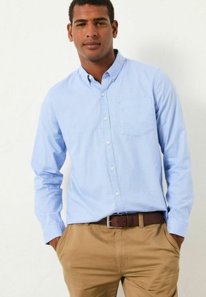 Šviesiai mėlynas ilgas rankovės marškiniai su viena krūtine kišene, derinami su khaki kelnėmis ir rudu diržu. Lygus audinio tekstūra.