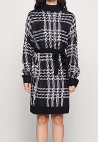 Abito lavorato a maglia a scacchi bianco e nero con maniche lunghe, polsini a coste e una cintura abbinata da annodare in vita. Texture morbida e vestibilità comoda.
