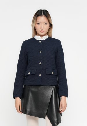 Donna che indossa un giacca blu navy con texture e bottoni sopra una blusa bianca, abbinata a una gonna in pelle nera asimmetrica e collant leggeri.