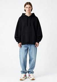 Joven de pie vistiendo una sudadera negra con capucha, jeans claros holgados y zapatillas blancas con acentos rojos y azules sobre un fondo blanco.