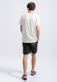 Witte t-shirt met korte mouwen in een ontspannen pasvorm, gecombineerd met zwarte cargoshorts met olijfgroene accenten en meerdere zakken. Sandalen maken de look af.