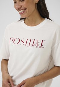 Bílá bavlněná tričko s krátkým rukávem, na kterém je červený nápis "POZITIVNÍ vibrace". Látka vypadá měkce a pohodlně.