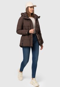 Bruine pufferjack met capuchon, voorzien van een ritssluiting en knopen, zijzakken en een geborduurd ontwerp. Gedragen met blauwe jeans en sneakers.