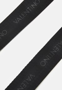 Černý látkový pásek s vyraženým nápisem "Valentino". Texturovaný povrch s tkaným vzorem. Dva paralelní pásky, detailní pohled.